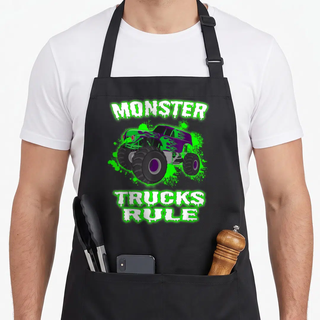 Rule truck apron - Purple monster truck with green splatter and white dripping text reading MONSTER TRUCKS RULE — shown on a black cotton canvas apron against a white background