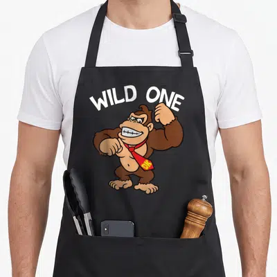 Wild donkey apron - Brown cartoon gorilla wearing red tie under white arched WILD ONE text — shown on a black cotton canvas apron against a white background