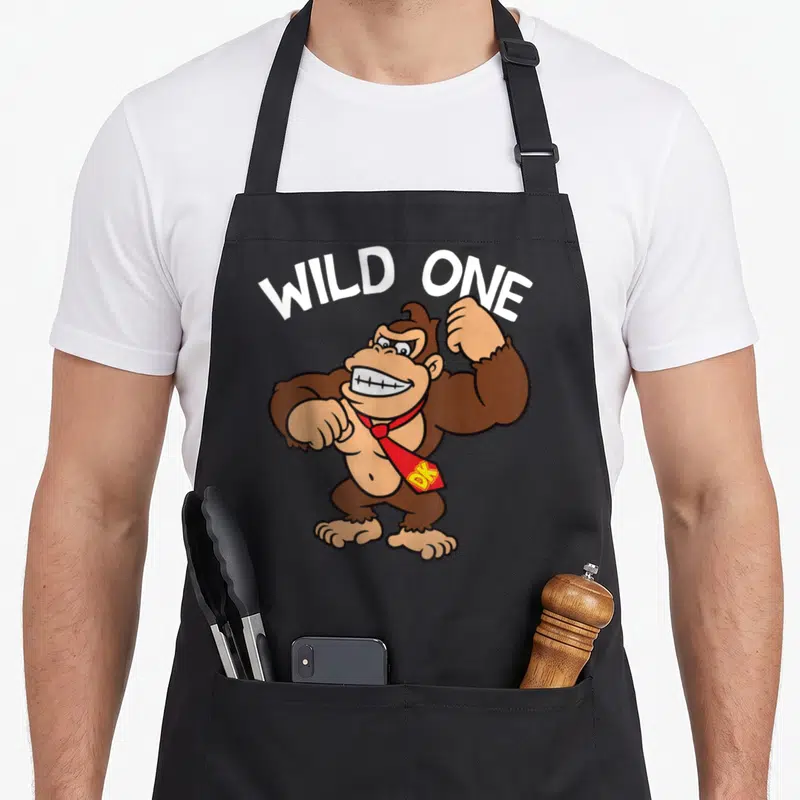 Wild donkey apron - Brown cartoon gorilla wearing red tie under white arched WILD ONE text — shown on a black cotton canvas apron against a white background