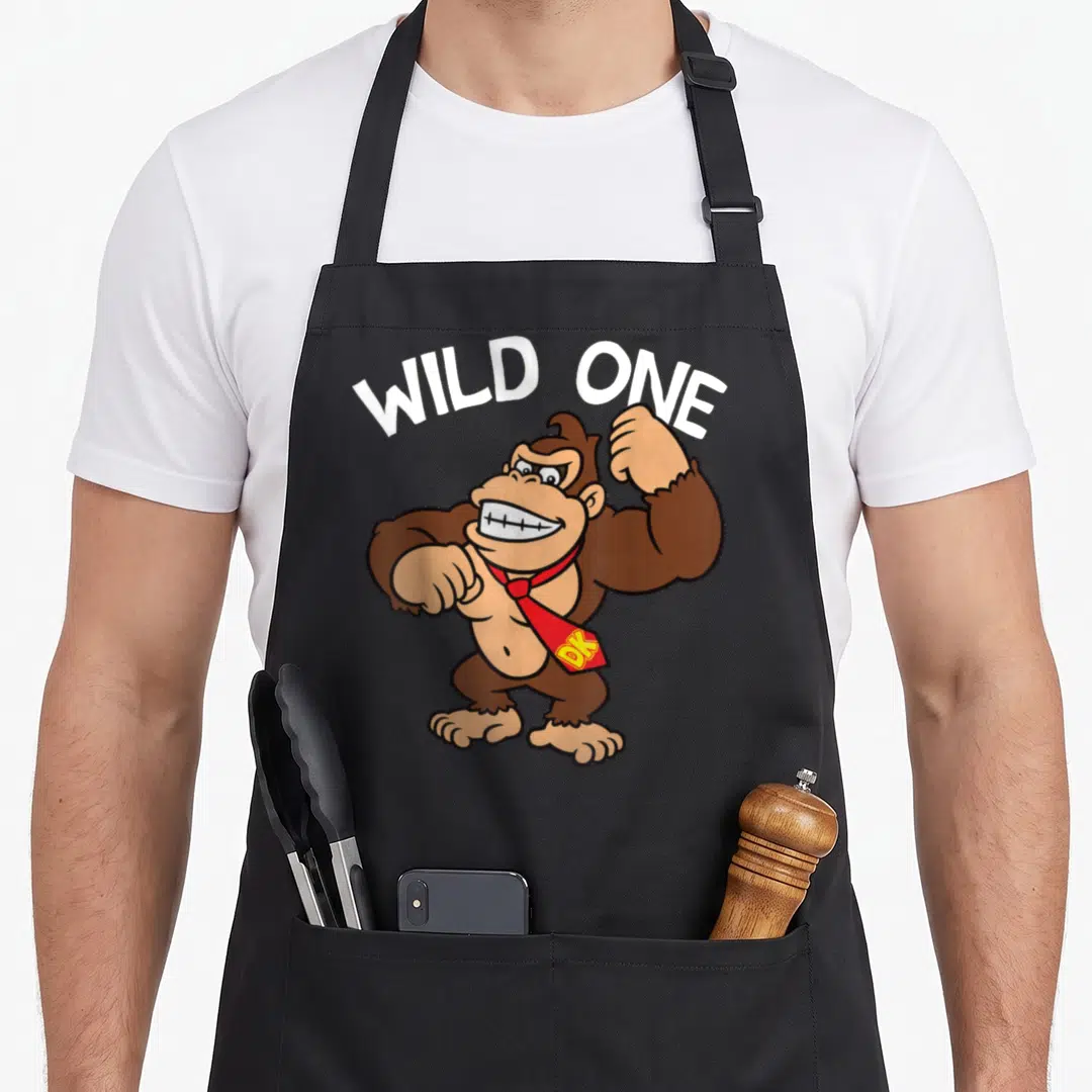 Wild donkey apron - Brown cartoon gorilla wearing red tie under white arched WILD ONE text — shown on a black cotton canvas apron against a white background