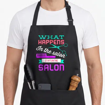 Hairstyling hairstylist apron - Stacked text reading WHAT HAPPENS In the salon STAYSINTHE SALON with teal scissors — shown on a black cotton canvas apron against a white background