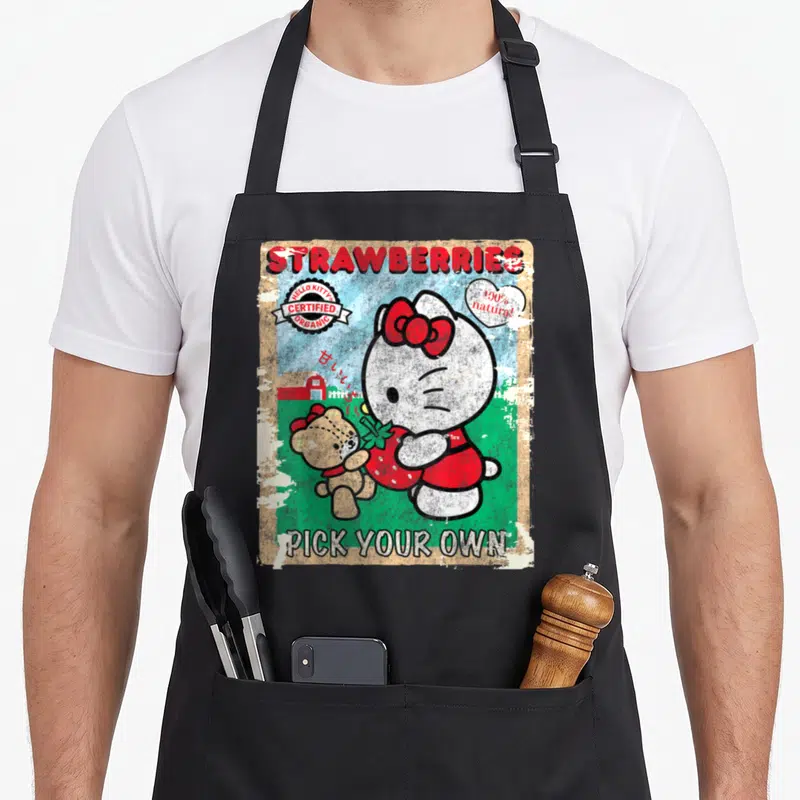 Picking strawberry apron - Hello Kitty and teddy bear holding red strawberry with barn, clouds, and distressed vintage — shown on a black cotton canvas apron against a white background
