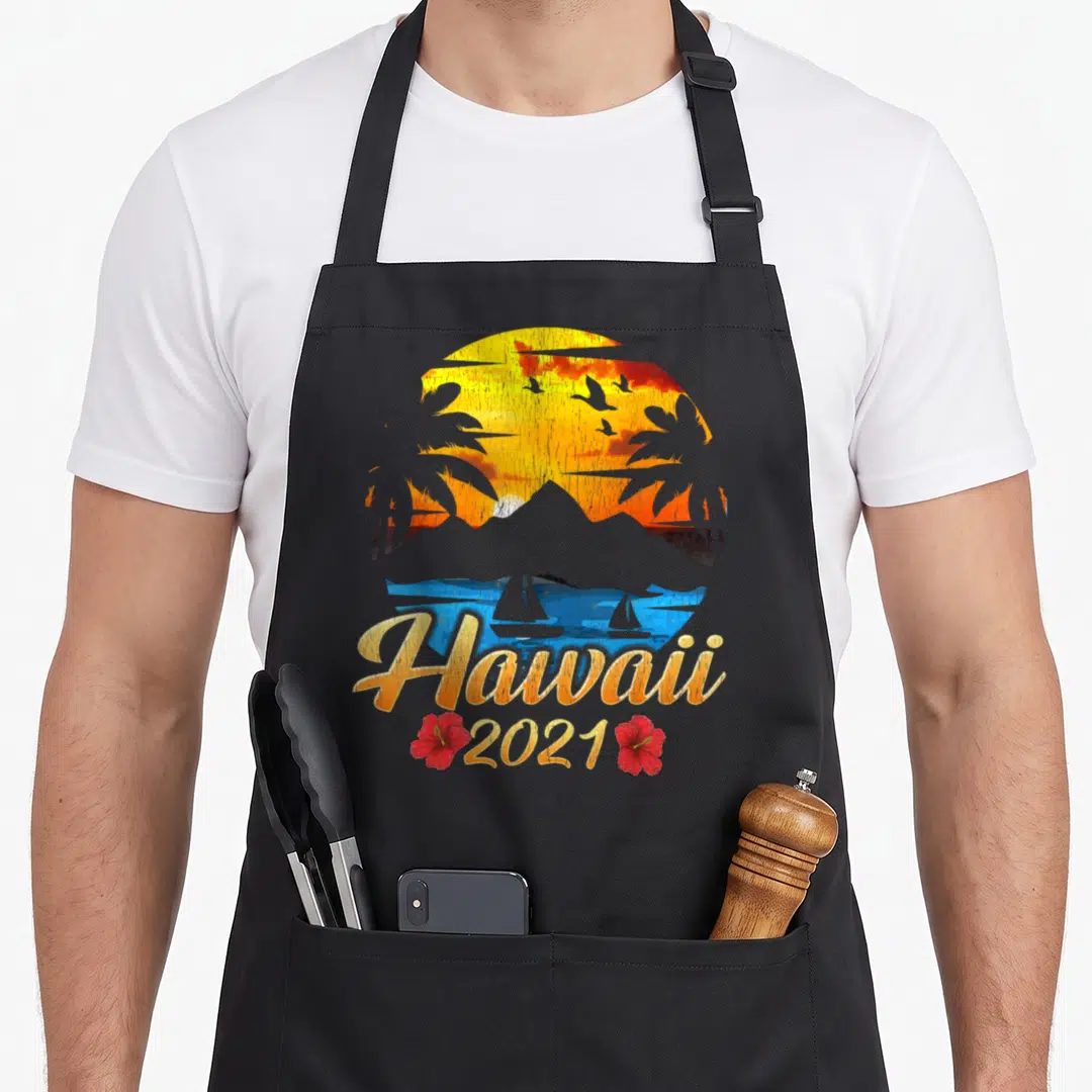 Family hawaiian apron - Retro circular sunset with palm trees, sailboats, Hawaii 2021 text, and two red hibiscus flowers — shown on a black cotton canvas apron against a white background