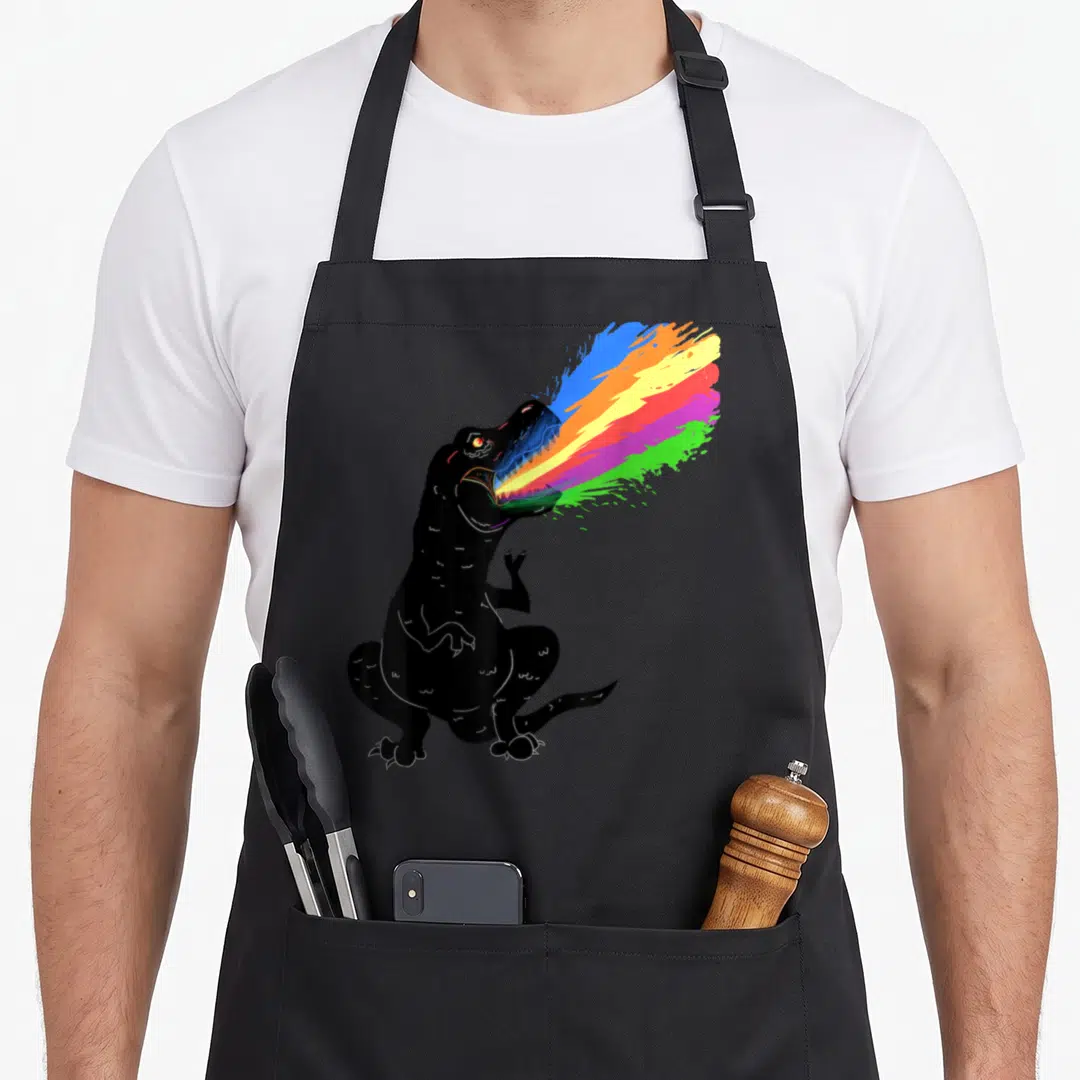 Tyrannosaurus shooting apron - Sitting black cartoon dinosaur breathing a rainbow stream of blue orange yellow pink — shown on a black cotton canvas apron against a white background