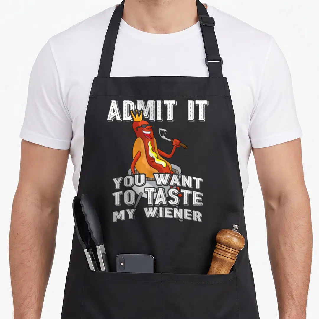 Admit sausage apron - Crowned cartoon hot dog on grill with ADMIT IT YOU WANT TO TASTE MY WIENER text — shown on a black cotton canvas apron against a white background