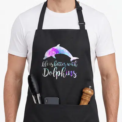 Better dolphin lover apron - Gradient dolphin silhouette above Life is Better With Dolphins script in purple and blue — shown on a black cotton canvas apron against a white background