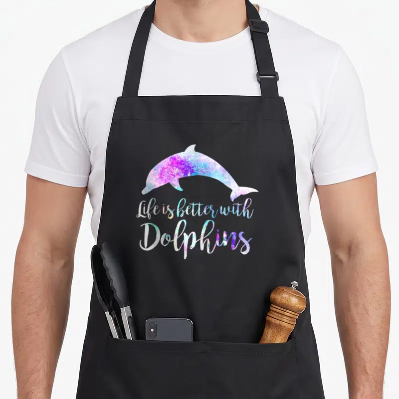 Better dolphin lover apron - Gradient dolphin silhouette above Life is Better With Dolphins script in purple and blue — shown on a black cotton canvas apron against a white background