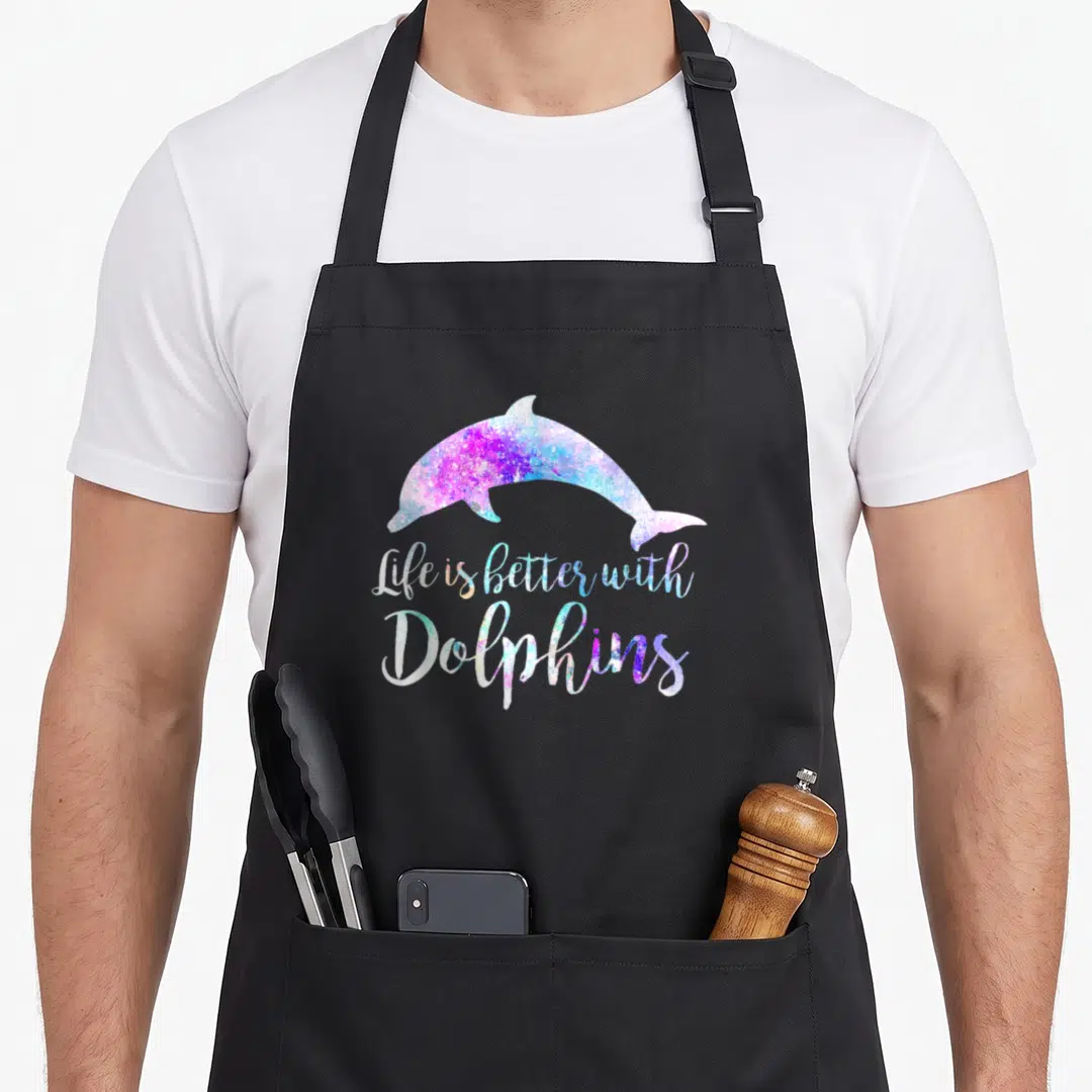Better dolphin lover apron - Gradient dolphin silhouette above Life is Better With Dolphins script in purple and blue — shown on a black cotton canvas apron against a white background