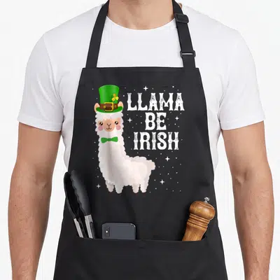 Llama st patrick day aprons - Cartoon white llama in green hat and bow tie beside stacked text LLAMA BE IRISH on grey — shown on a black cotton canvas apron against a white background