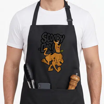 Vintage decade apron - Cartoon brown dog overlapping large grey Scooby, Doo! text with yellow tag detail — shown on a black cotton canvas apron against a white background