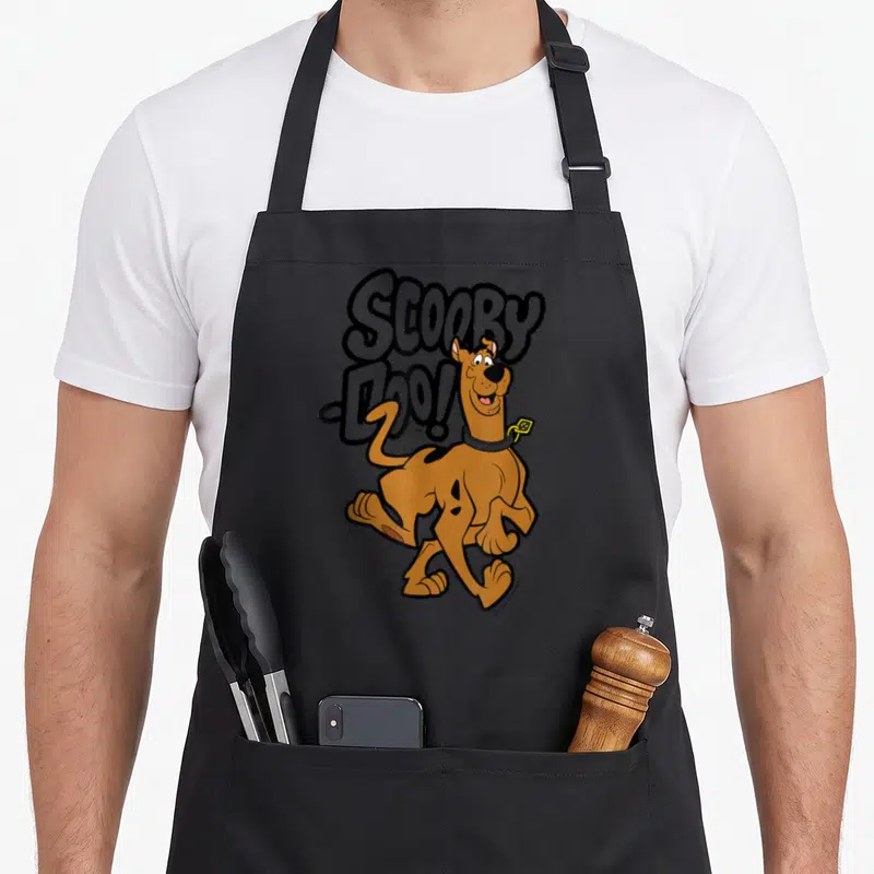 Vintage decade apron - Cartoon brown dog overlapping large grey Scooby, Doo! text with yellow tag detail — shown on a black cotton canvas apron against a white background