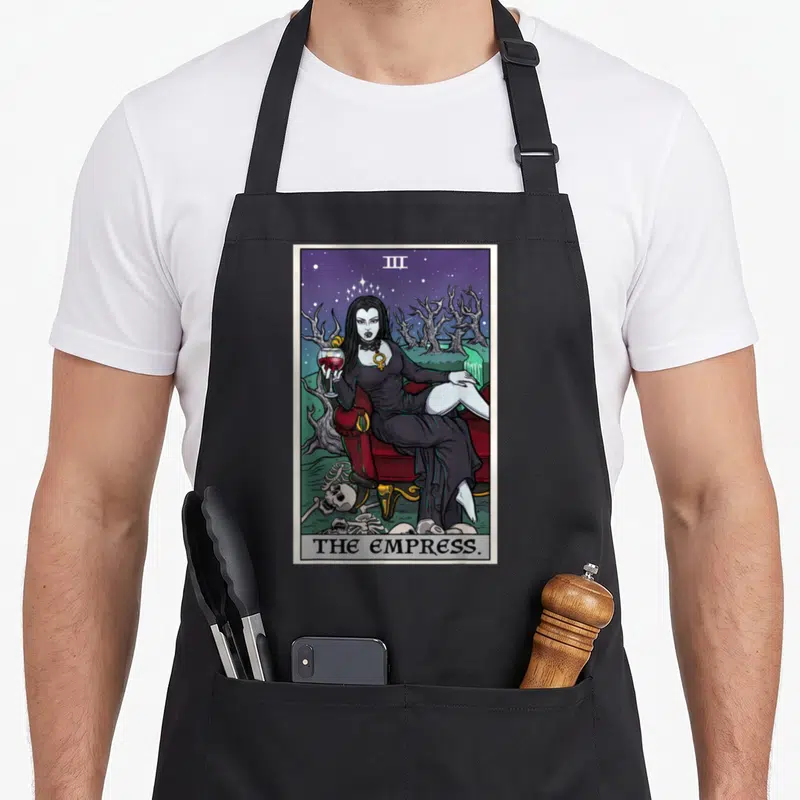 Empress halloween witch apron - Gothic Empress tarot card woman sitting on red couch with skeletons and starry sky — shown on a black cotton canvas apron against a white background