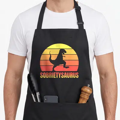 Sobriety t-rex apron - Grey T-Rex silhouette on striped yellow orange red sun with SOBIETYSAURUS text below — shown on a black cotton canvas apron against a white background