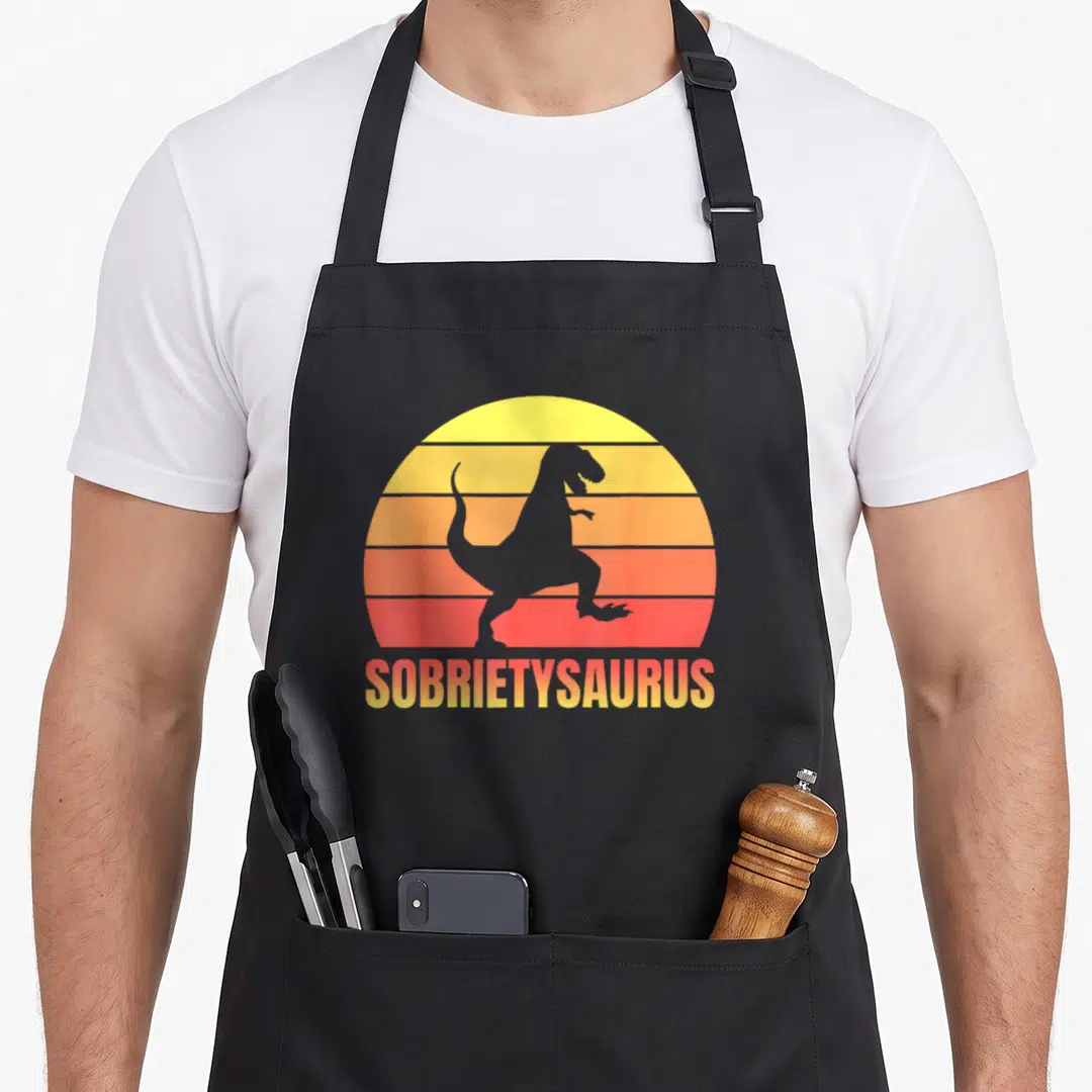 Sobriety t-rex apron - Grey T-Rex silhouette on striped yellow orange red sun with SOBIETYSAURUS text below — shown on a black cotton canvas apron against a white background