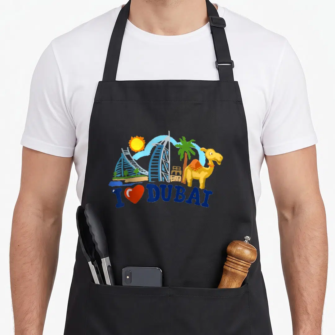 Skyscraper arab apron - Cartoon Dubai skyline with Burj Al Arab, camel, and I Love Dubai text in blue, yellow, red — shown on a black cotton canvas apron against a white background