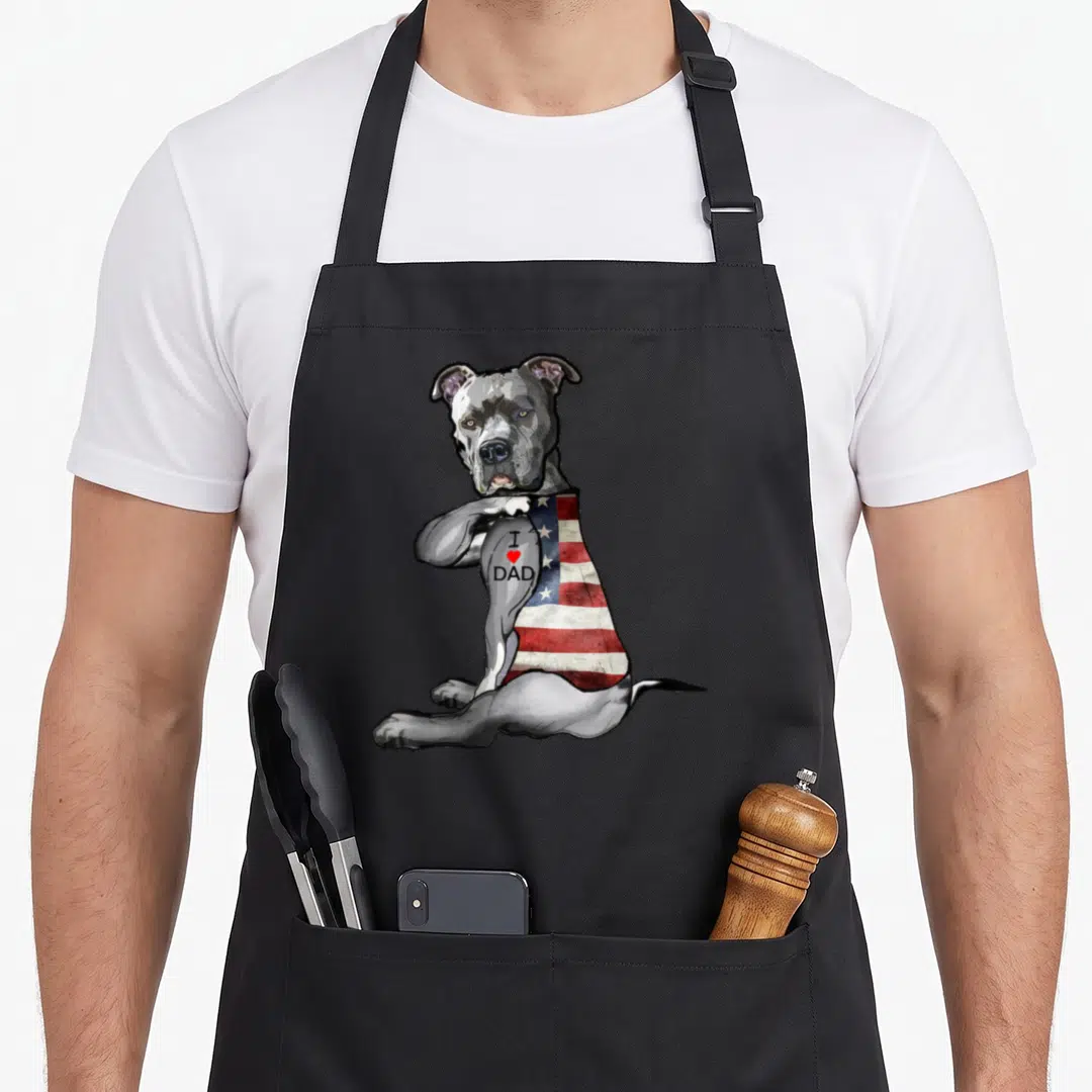 Pitbull tattoo aprons - Grey dog sitting with American flag back and I love dad tattoo on arm — shown on a black cotton canvas apron against a white background