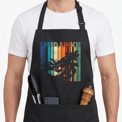 Distressed cross apron - SPEEDJUNKIE text above grey motorcycle silhouette on blue, teal, yellow, orange, red — shown on a black cotton canvas apron against a white background