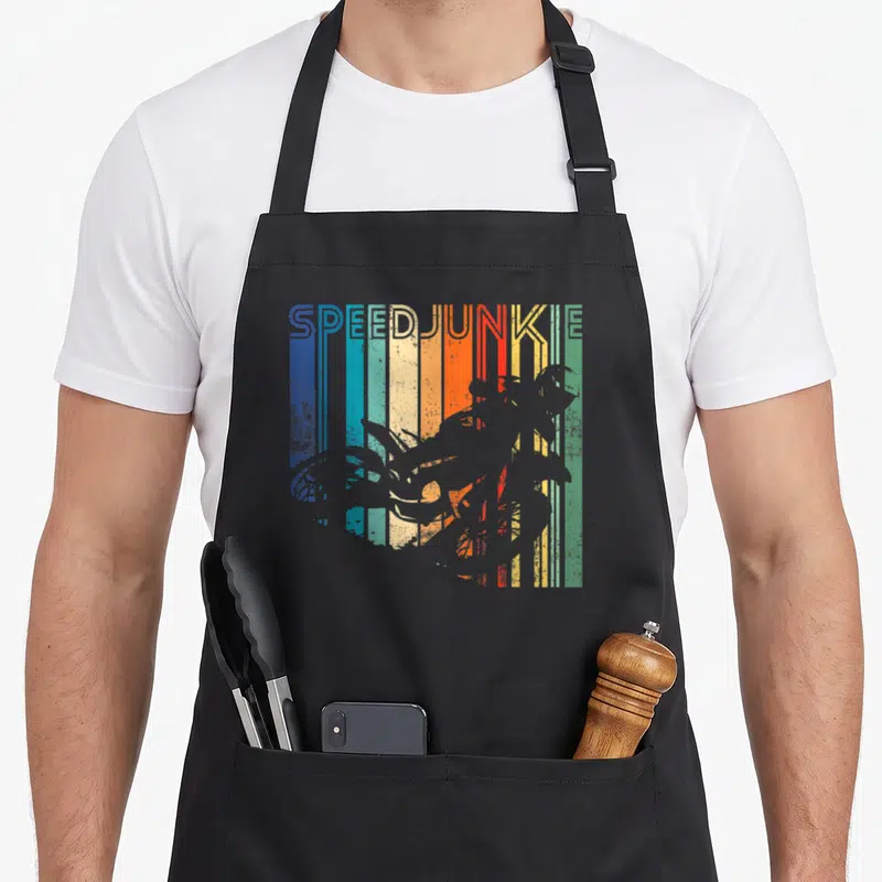 Distressed cross apron - SPEEDJUNKIE text above grey motorcycle silhouette on blue, teal, yellow, orange, red — shown on a black cotton canvas apron against a white background