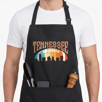 Skyline football apron - Tennessee text arched over a colorful skyline football in distressed orange, white, blue — shown on a black cotton canvas apron against a white background