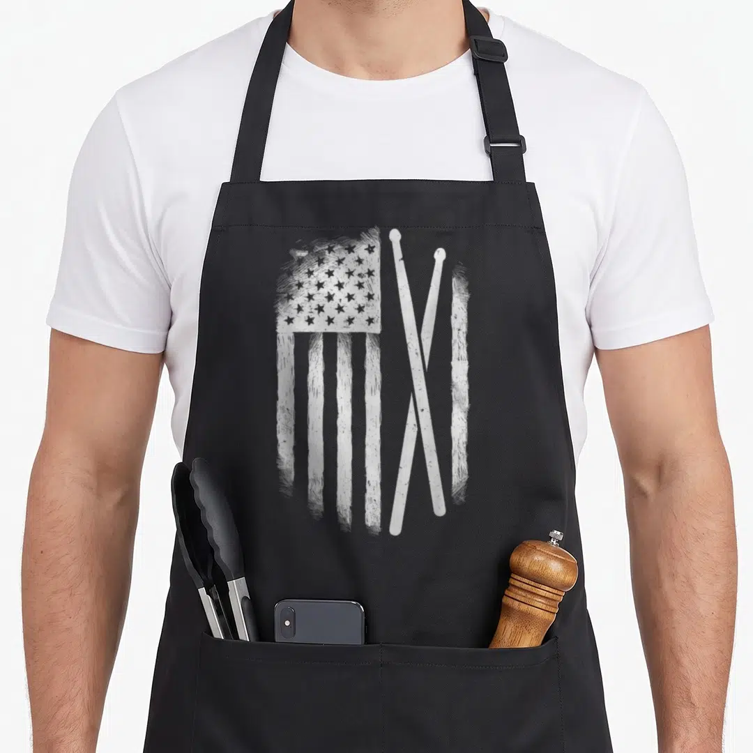 Drumstick american flag apron - Distressed white USA flag and two crossed drumsticks graphic — shown on a black cotton canvas apron against a white background