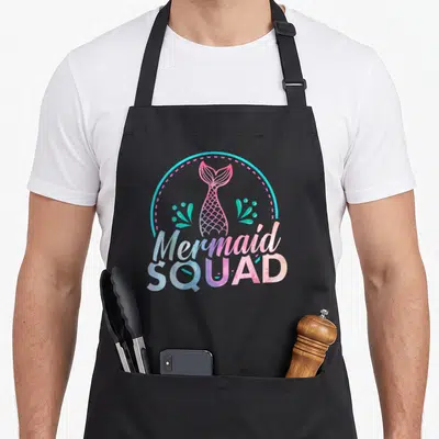 Squad birthday apron - Watercolor mermaid tail in pink and purple inside cyan circle with seaweed and text — shown on a black cotton canvas apron against a white background