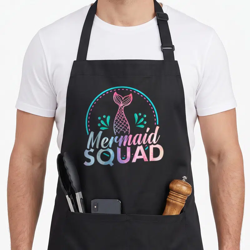 Squad birthday apron - Watercolor mermaid tail in pink and purple inside cyan circle with seaweed and text — shown on a black cotton canvas apron against a white background
