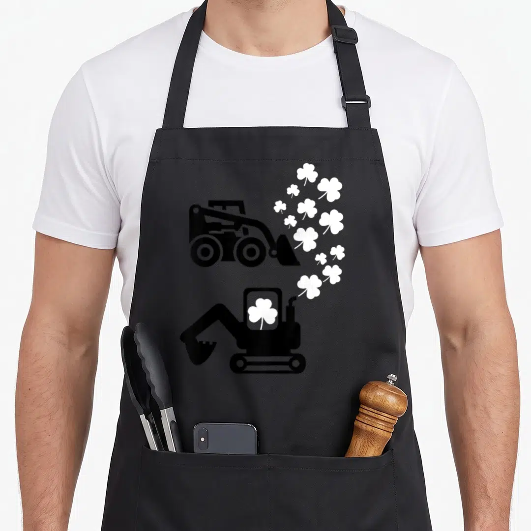 Patrick truck apron - Black skid steer and excavator with white shamrocks floating from bucket and exhaust pipe — shown on a black cotton canvas apron against a white background