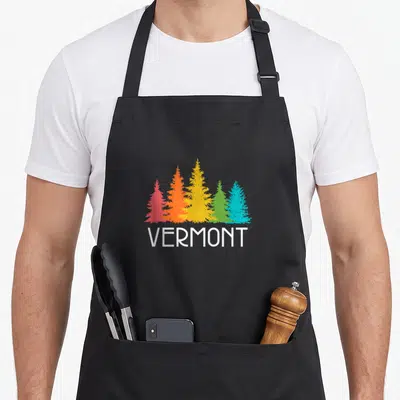 Cute vermont apron - Five colorful pine trees in red orange yellow green blue white above white VERMONT text — shown on a black cotton canvas apron against a white background