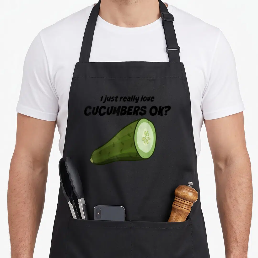 Freaking honestly apron - Black text reading I just really love CUCUMBERS OK above a green cartoon cucumber slice — shown on a black cotton canvas apron against a white background