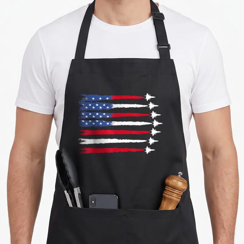 Fighter 4th of july apron - Seven white jets leaving distressed smoke trails forming a US flag pattern in red white — shown on a black cotton canvas apron against a white background