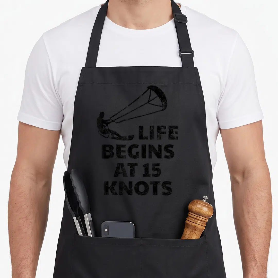 Kiteboarding joke apron - Black kitesurfer silhouette with LIFE BEGINS AT 15 KNOTS stacked distressed text — shown on a black cotton canvas apron against a white background