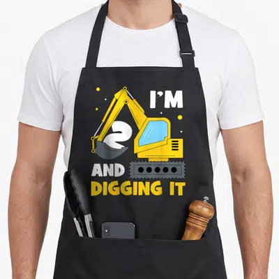 Digging 2nd apron - Yellow excavator over white number 2 with text I'M 2 AND DIGGING IT — shown on a black cotton canvas apron against a white background