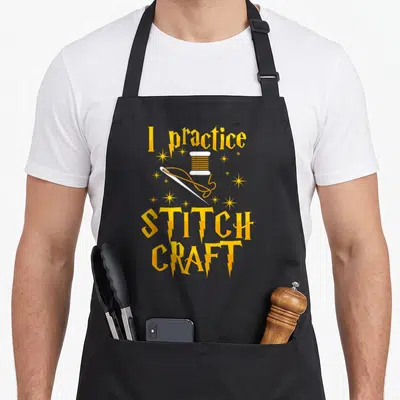 Practice cross stitch aprons - I practice STITCH CRAFT text with yellow spool needle and stars — shown on a black cotton canvas apron against a white background