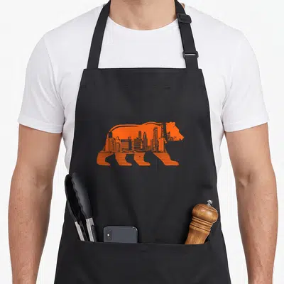 Downtown skyline illinois chicago apron - Orange bear silhouette with city skyline integrated inside, formed — shown on a black cotton canvas apron against a white background