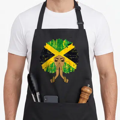 Jamaican queen apron - Cartoon woman with praying hands and afro filled with green yellow black and brown flag pattern — shown on a black cotton canvas apron against a white background