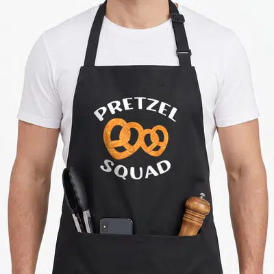 Squad soft pretzel apron - Three interlocking golden brown pretzels with white PRETZEL SQUAD text arched above and below — shown on a black cotton canvas apron against a white background