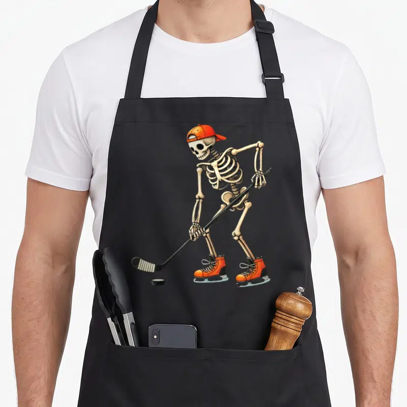Skeleton ice halloween apron - Cartoon skeleton in orange skates and cap holding a hockey stick over a black puck — shown on a black cotton canvas apron against a white background