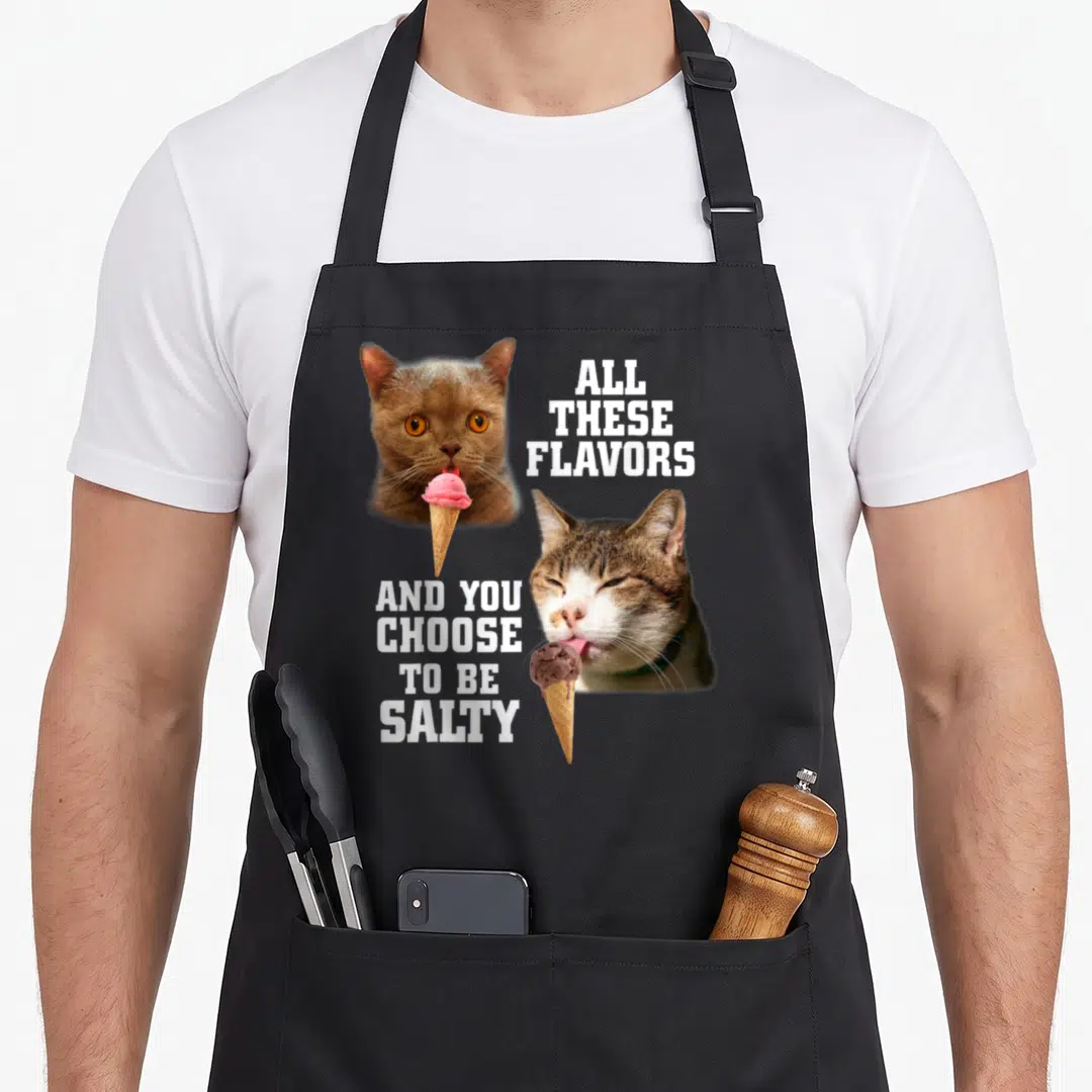 These cat apron - Two cats eating pink and brown ice cream with white text ALL THESE FLAVORS AND YOU CHOOSE TO BE SALTY — shown on a black cotton canvas apron against a white background