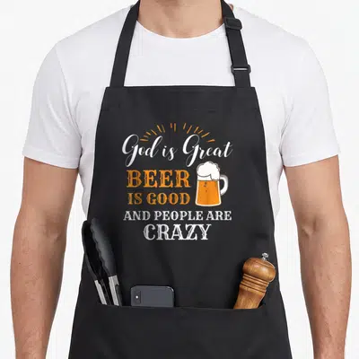 Product beer apron - Stacked white and orange text reading God is Great Beer Is Good next to a beer mug illustration — shown on a black cotton canvas apron against a white background