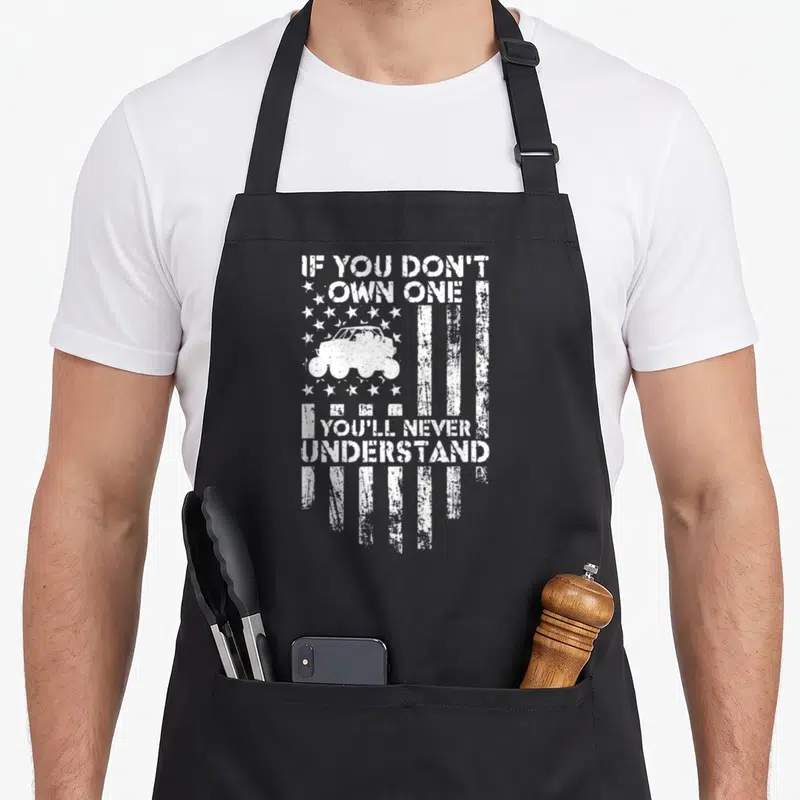 Offroading youll never apron - White distressed US flag graphic with monster truck and text IF YOU DON'T OWN ONE — shown on a black cotton canvas apron against a white background