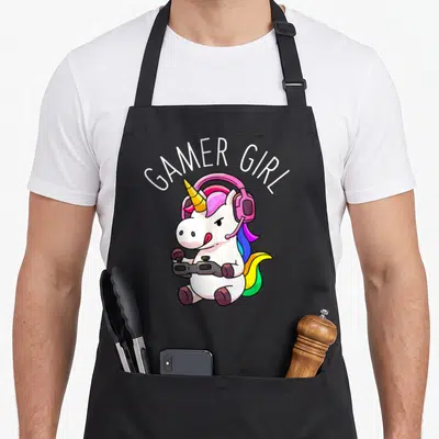 Gamer unicorn apron - Cartoon white unicorn with pink headphones holding black controller under arched GAMER GIRL text — shown on a black cotton canvas apron against a white background