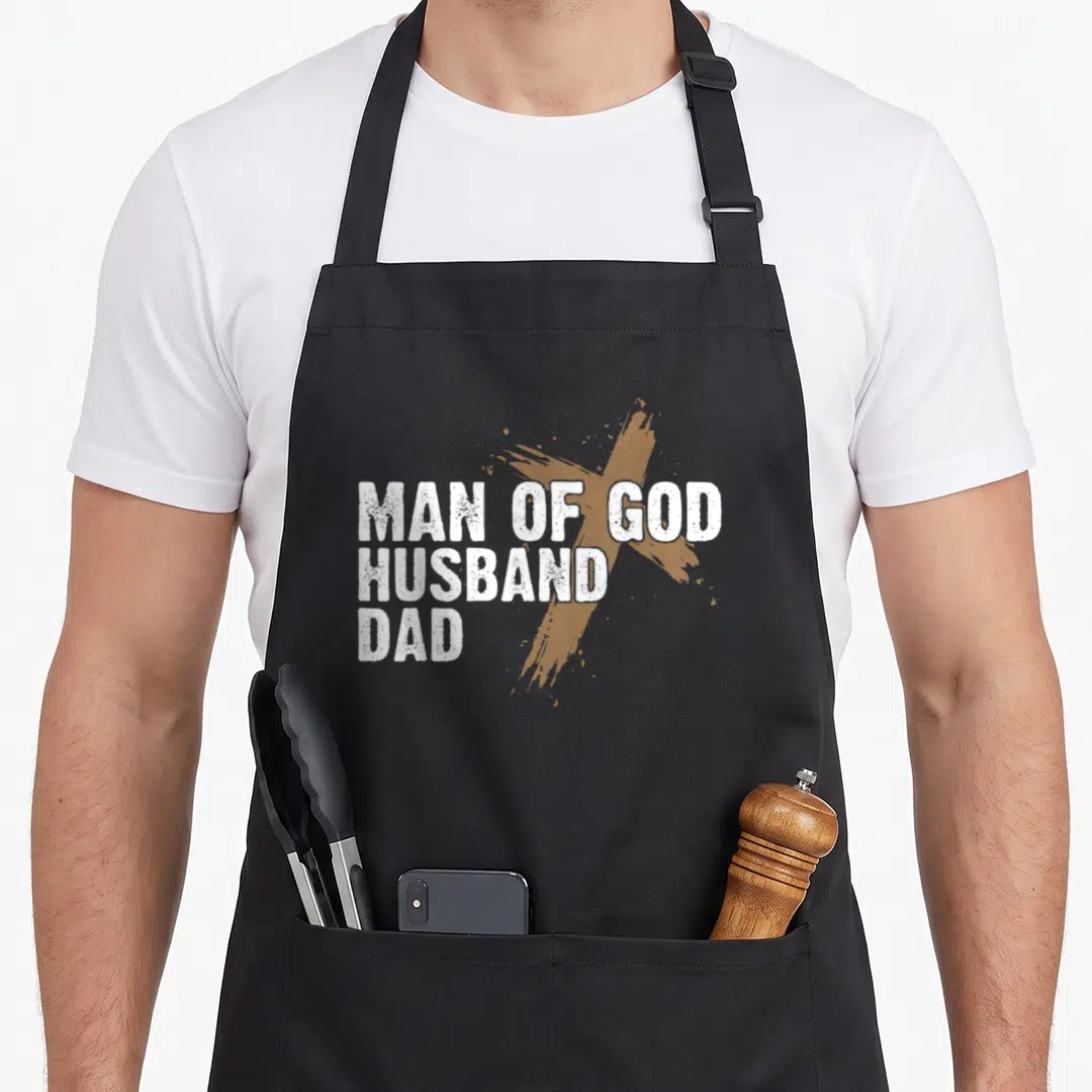 Faith father day apron - MAN OF GOD HUSBAND DAD white text over tan brush-stroke cross, distressed style — shown on a black cotton canvas apron against a white background