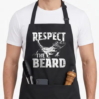 Respect dragon apron - White distressed bearded dragon illustration between text RESPECT THE BEARD — shown on a black cotton canvas apron against a white background