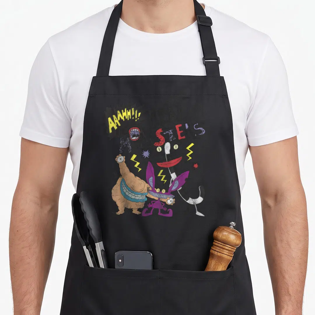 Aaahh friends apron - Three monsters below AAAHH!! REAL MONSTERS text with yellow purple orange black lightning bolts — shown on a black cotton canvas apron against a white background