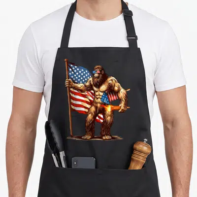 Bigfoot firework 4th of july apron - Brown sasquatch holding US flag and lit torch wearing patriotic sunglasses — shown on a black cotton canvas apron against a white background