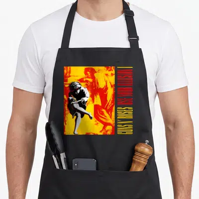 Use gun apron - Retro man figure with vertical Guns N Roses Use Your Illusion I text in yellow red white black — shown on a black cotton canvas apron against a white background