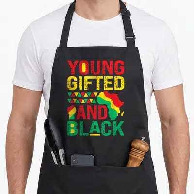 Young nourjoy apron - Young Gifted And Black text with Africa map and raised fist in red yellow green distressed style — shown on a black cotton canvas apron against a white background