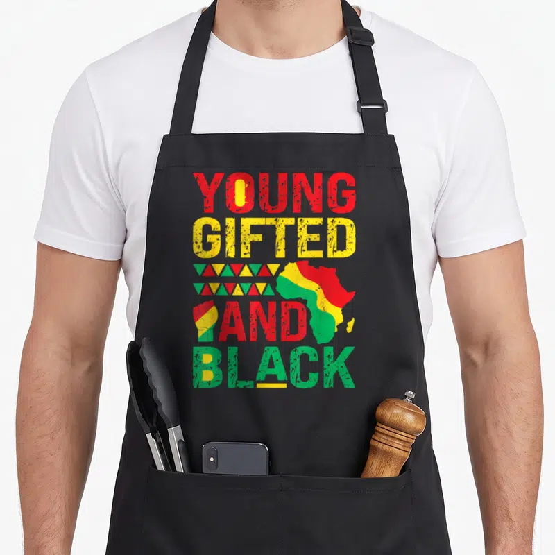 Young nourjoy apron - Young Gifted And Black text with Africa map and raised fist in red yellow green distressed style — shown on a black cotton canvas apron against a white background