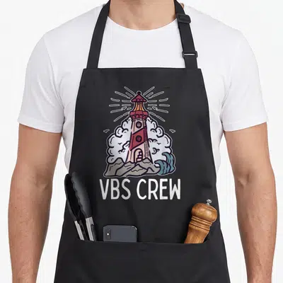 Vbs crew apron - Cartoon lighthouse with red stripes and VBS CREW text below surrounded by clouds and waves — shown on a black cotton canvas apron against a white background