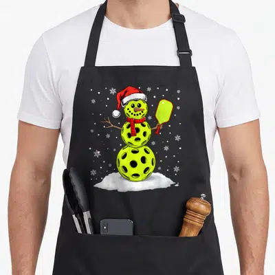 Hat snowman apron - Lime green pickleball snowman wearing red Santa hat and scarf holding a paddle on white snow — shown on a black cotton canvas apron against a white background